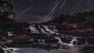 Vista noturna de uma cachoeira que escorre sobre degraus de pedra a poucos metros de altura em relação ao seu poço. Ao fundo, a estrelas criam rastros de luz devido à técnica fotográfica de longa exposição