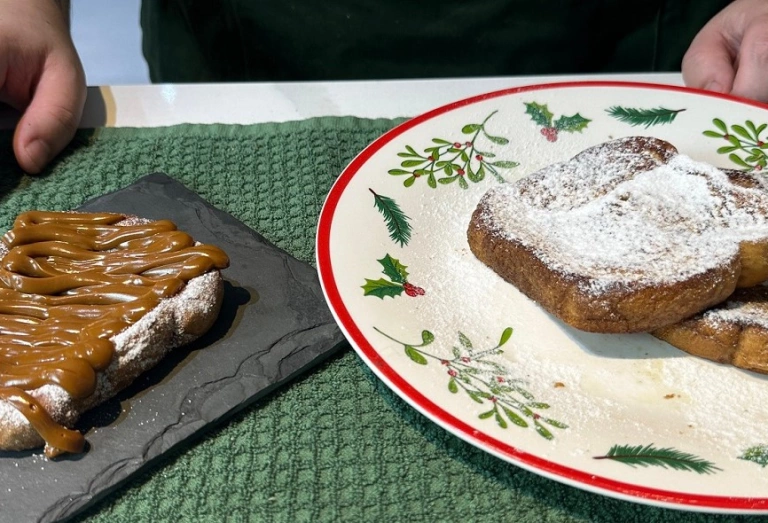 Um prato com decorações natalinas e uma pedra de ardósia servidas de fatias de pães. Uma, coberta de açúcar e doce de leite, outras duas, somente de açúcar. É conhecida como rabanada.