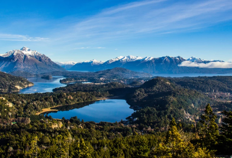 Vista aérea do Lago Nahuel Huapi, Bariloche, Argentina, com os picos nevados das montanhas ao fundo e trechos de floresta ao redor.