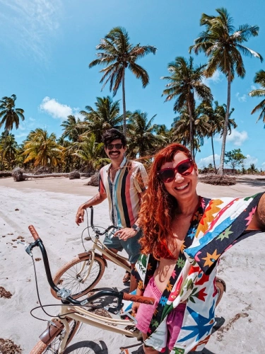 Uma mulher  e um homem a bordo de duas bicicletas na beira do mar com coqueiros ao fundo da paisagem.