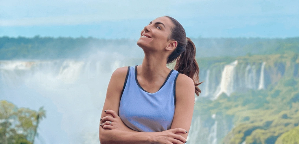Mulher de braços cruzados e encostada em uma grade sorri e olha emocionada para o céu. Ao fundo, as quedas d’água das Cataratas do Iguaçu.