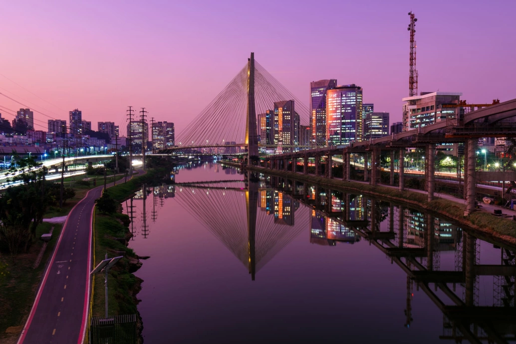 Vista da cidade de São Paulo ao anoitecer. No centro da foto está a Ponte Estaiada, cercada por grandes edifícios e refletindo sobre as águas do rio