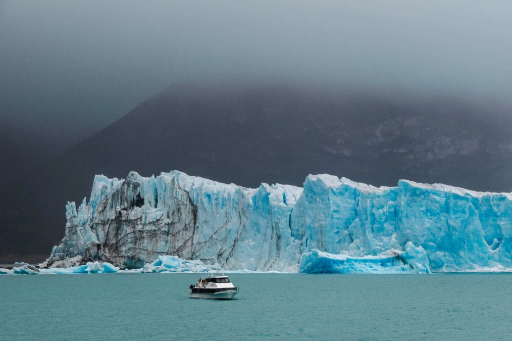 Se destacando da neblina ao fundo, surge a geleira Glaciar Perito Moreno, localizada no Parque Nacional Los Glaciares, Argentina. Aos seus pés, uma formação de água esverdeada onde um iate circula