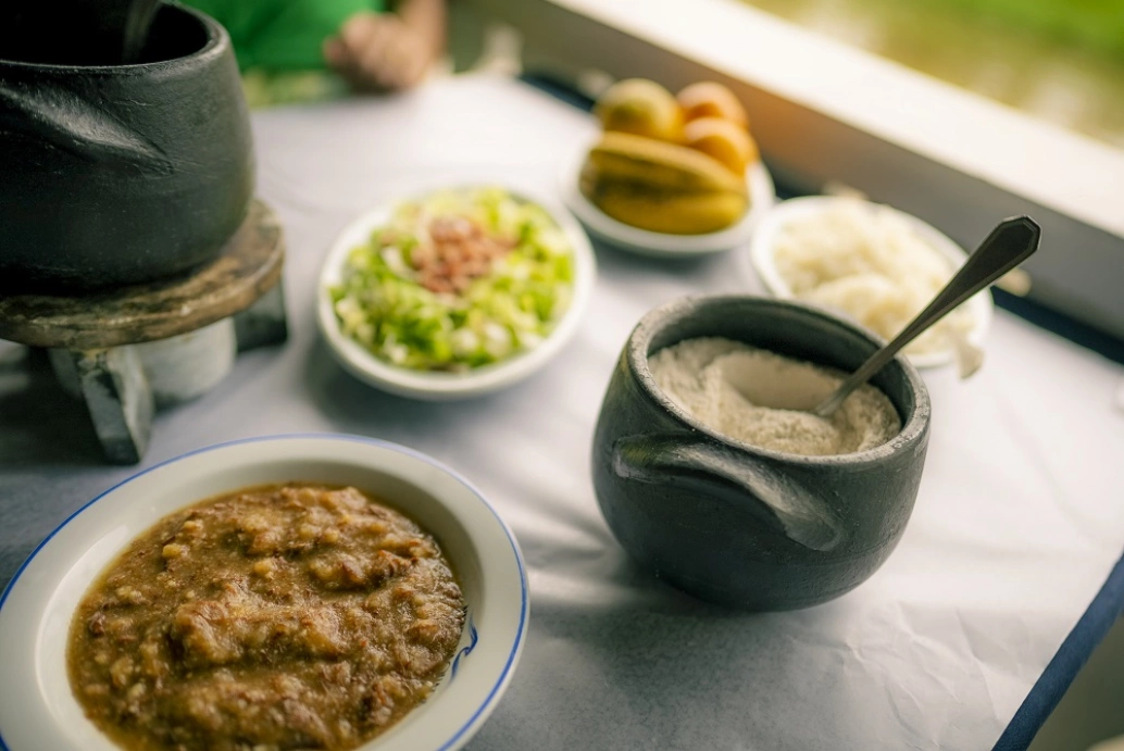 Em uma mesa com forro branco, uma panela de barro, um prato branco de borda azul servido de barreado e um recipiente menor de barro com farinha. Ao fundo, salada, arroz e frutas.