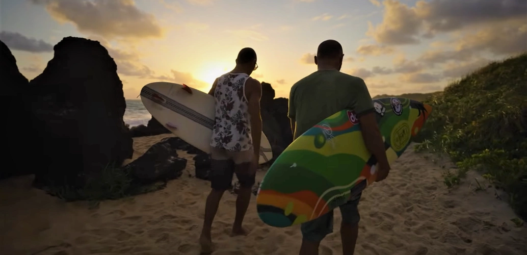Dois homens caminham em praia segurando pranchas de surfe