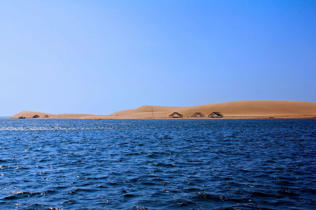 No plano frontal, águas do Delta Parnaíba. Ao fundo, três construções típicas de pescadores, e dunas de areia
