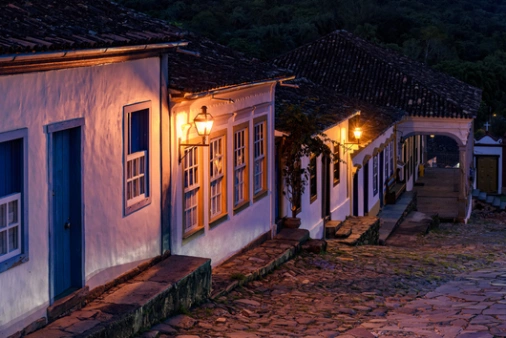 Vista noturna de rua de pedra com casas em arquitetura colonial, com luminárias externas acesas