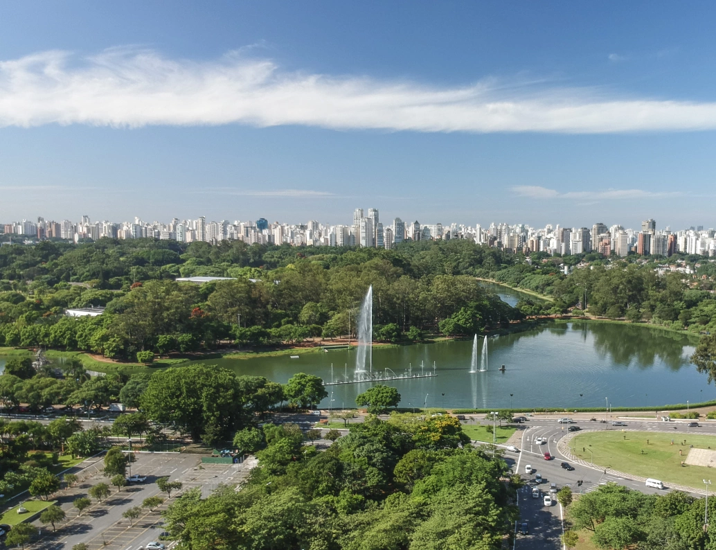 Vista aérea do Parque do Ibirapuera em São Paulo. Há um grande lago com fontes de água em meio à extensa área verde. Ao fundo, uma infinidade de edifícios