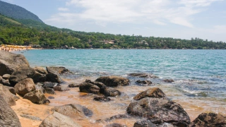 Vista ampla de um mar azul que se encontra com algumas pedras à beira-mar. Ao fundo, extensa área de vegetação nativa