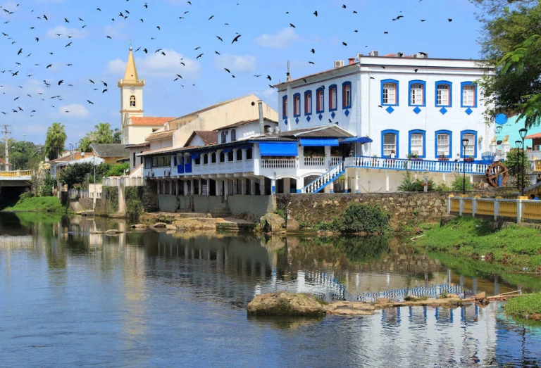 Dia ensolarado com revoada de pássaros sobre um rio que atravessa a cidade. Construções coloniais nas margens do rio em Morretes