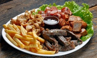 Um prato branco em uma mesa de madeira escura com batatas fritas, carne de boi, torresmo, carne de porco, linguiça e decorado com alface. Ao meio, um molho escuro.
