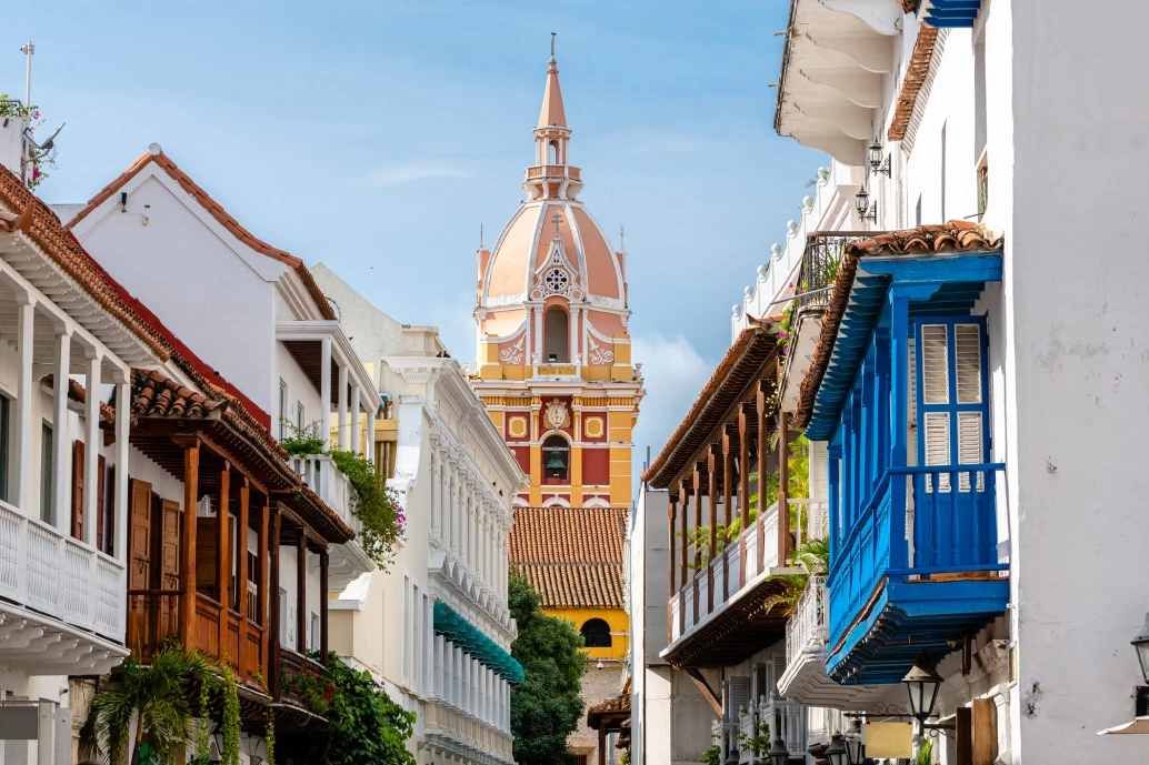 Entre sacadas de casas numa rua estreita de Cartagena, aparece o topo da catedral alaranjada ao final da rua, num dia claro. A igreja e as construções à sua volta possuem arquitetura colonial e cores vibrantes.