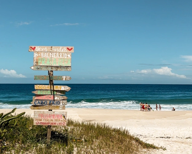 Placa de madeira em praia indicando as direções para Arraial do Cabo, Rio de Janeiro, Saquarema, Búzios e Cabo Frio. No topo da placa, há os dizeres “Bem vindos. Balneário Praia Seca”