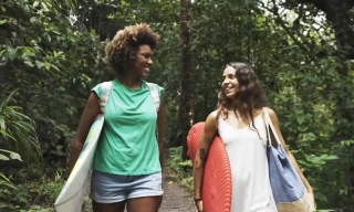 Duas mulheres sorrindo uma para a outra carregam pranchas de surfe debaixo do braço descendo uma trilha cercada por árvores
