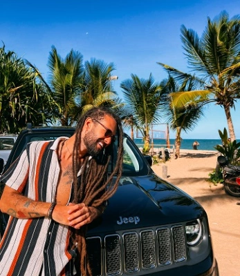 Músico Gabriel Elias se apoiando em dianteira de carro próximo à praia
