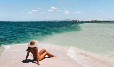 Mulher sentada em praia de areias claras avistando o verde mar e céu azul.