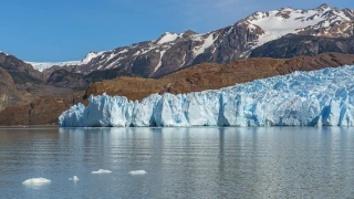 Enorme geleira de coloração azul na beira de um lago. Ao fundo, montanhas escuras com neve no topo