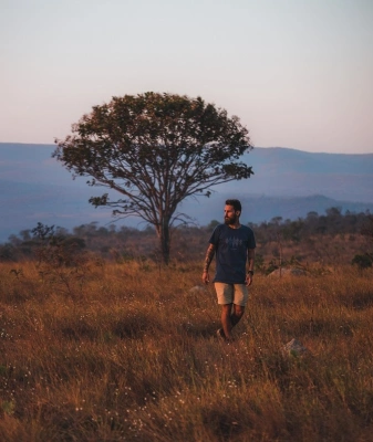 Homem caminha em espaço natural, com pasto de cor dourada, árvores e montanhas ao fundo.