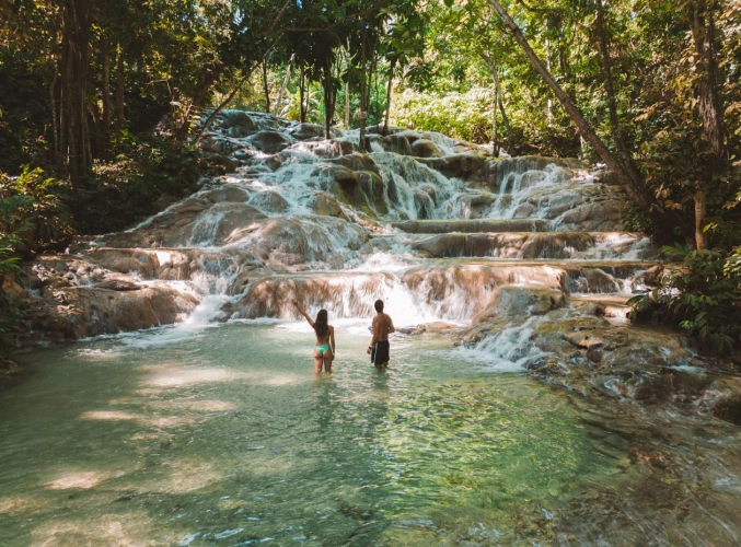 Mulher e homem de costas com meios corpo no poço da cachoeira enquanto apreciam a queda d’água que escorre em meio às pedras
