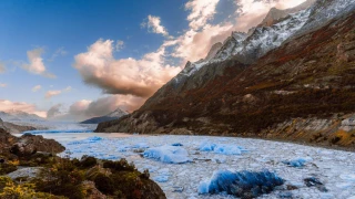 Lago com placas de gelo em sua superfície, em meio à paisagem da Patagônia chilena. Há relevos com vegetação nas margens, e montanhas nevadas ao fundo