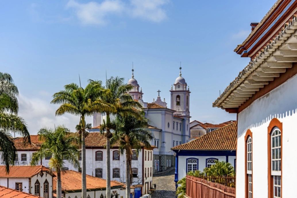 Construções coloniais, com destaque para uma igreja, e coqueiros em rua de pedra de uma cidade histórica em Minas Gerais