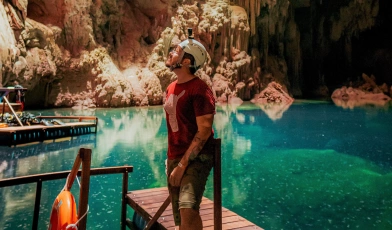 Homem posa para foto em gruta. Se destacam o verde cristalino da lagoa e as texturas das paredes.