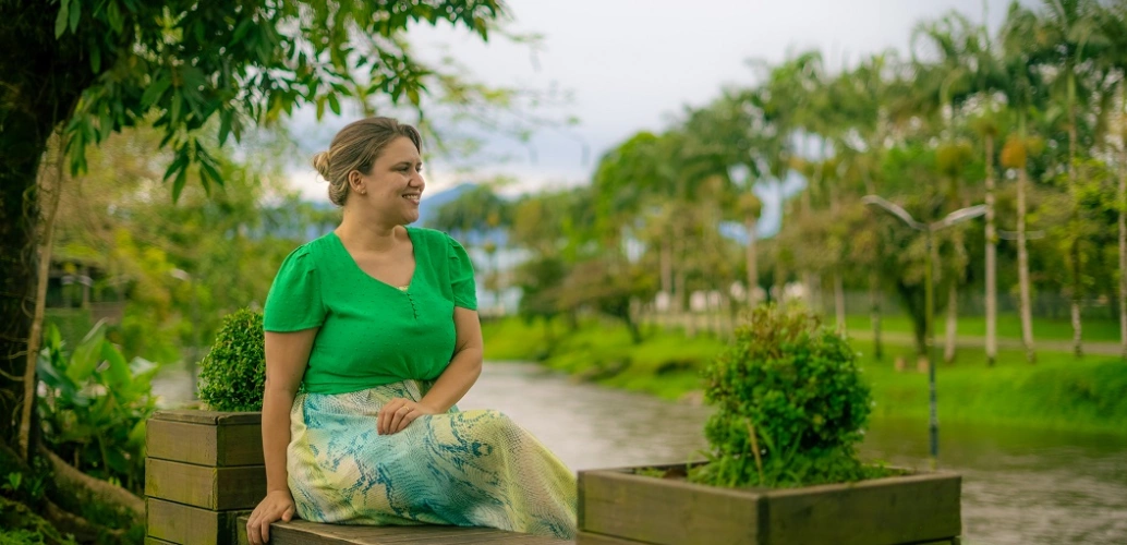 Mulher sorridente aprecia a vista sentada em banco de madeira em espaço aberto e arborizado durante o dia. O vibrante verde natural se destaca na imagem combinando com a roupa da mulher.