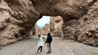 Casal de mãos dadas em meio a um cânion entre duas montanhas rochosas. O homem observa a ponte natural formada pelas pedras e a mulher olha para a câmera.