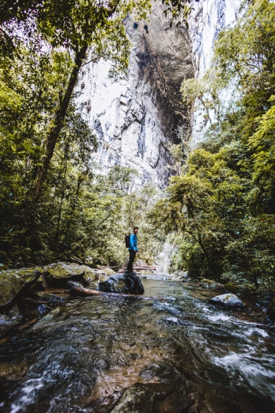 Homem em pé em cima de uma pedra em um rio, olhando para a câmera. Vegetação ao redor e enorme formação rochosa ao fundo