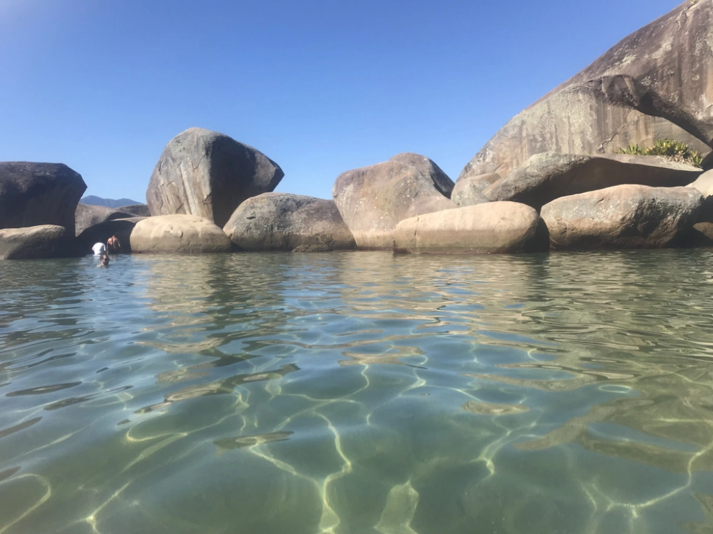 Destaque para água verde-esmeralda e algumas rochas formando uma barreira na piscina natural. Ao fundo, céu azul com poucas nuvens