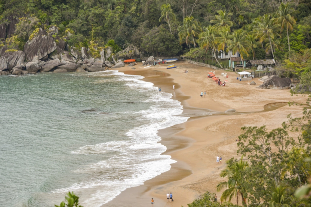 Praia vista pela lateral. Ondas invadem a faixa de areia onde estão alguns turistas. Ao redor, grandes pedras e densa vegetação nativa