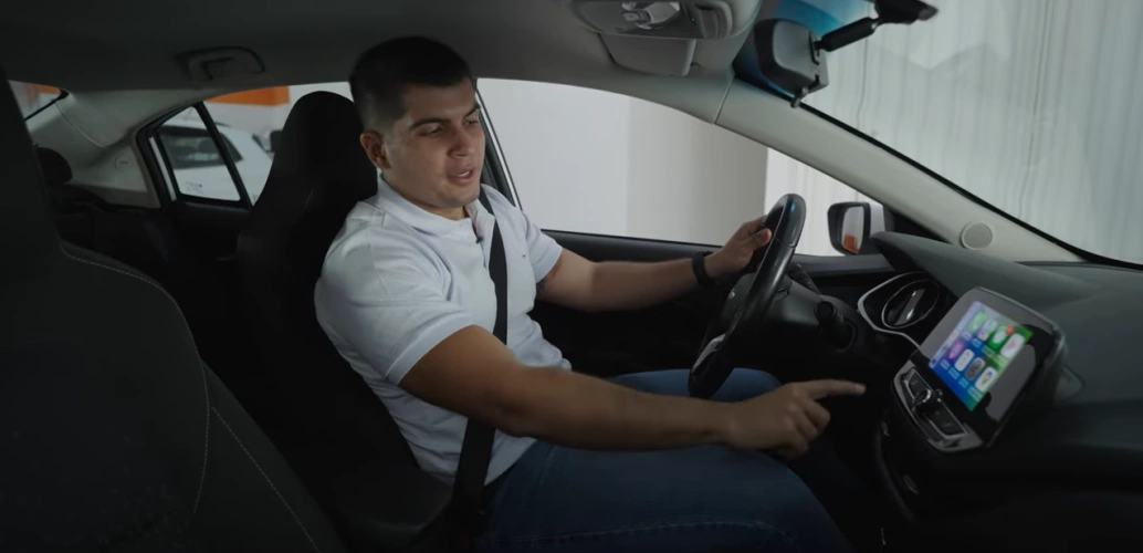 O influenciador Xenão está sentado no banco do motorista de um carro, com a mão esquerda ao volante e a outra apontando para a central multimídia.