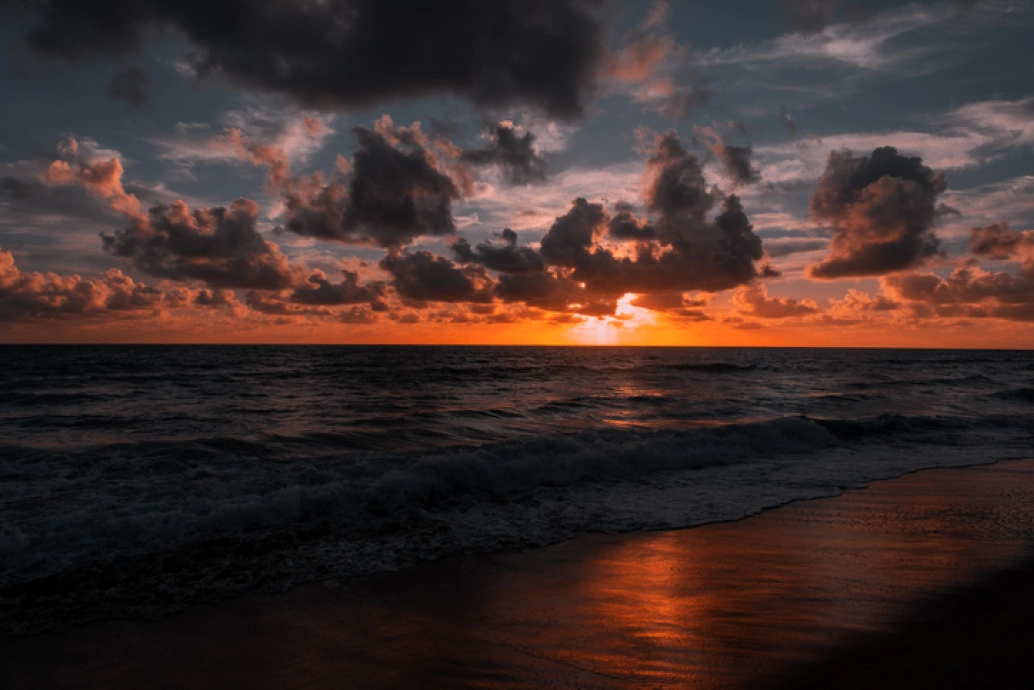 Pôr do sol cria silhueta das nuvens, formando um céu degradê do azul para o laranja. A luz do sol reflete no mar e na areia