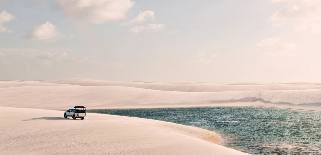 Cenário de um dia ensolarado com um carro passando por dunas que cercam um lago