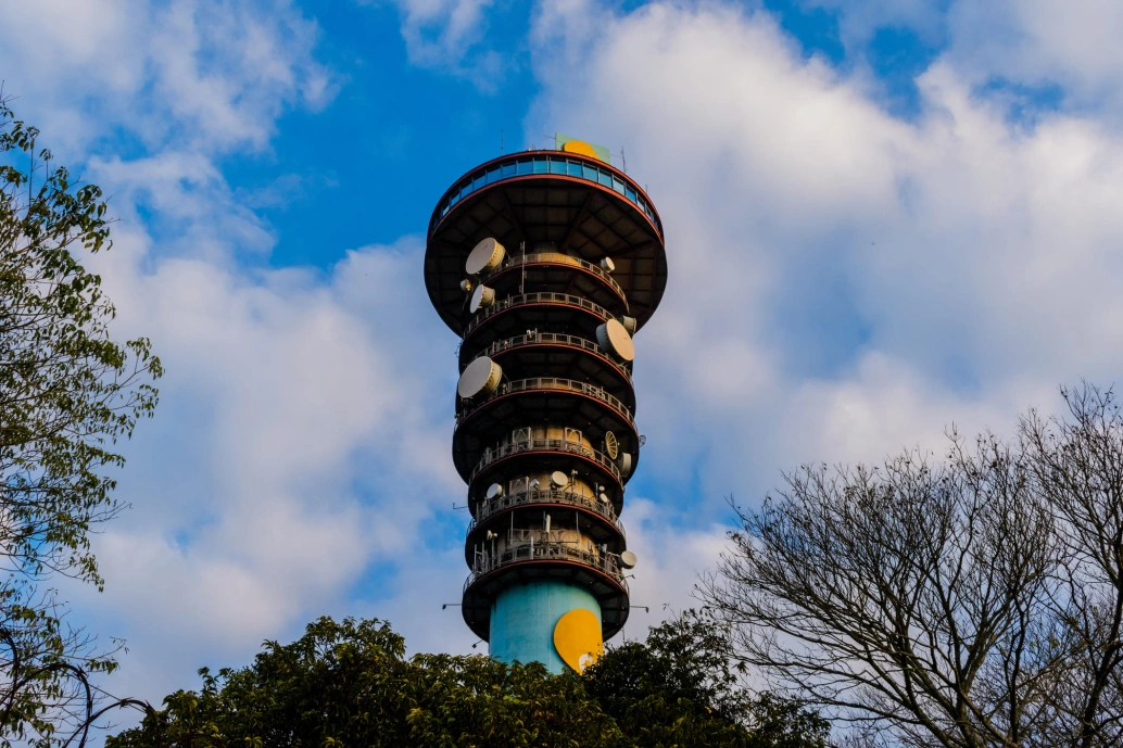 Topo de uma torre com mais de 100 metros de altura. Céu azul com algumas nuvens ao fundo e, ao redor da torre, aparecem copas e galhos de árvores