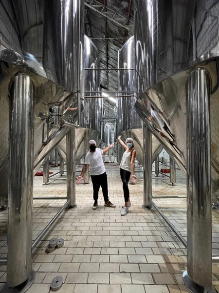 Um casal de máscaras e toucas visitando a fábrica de uma cervejaria em Santa Catarina, Brasil. Os dois estão entre os barris de alumínio, fazendo pose para a foto.