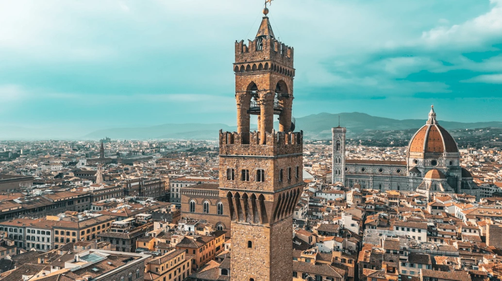 Vista aérea e panorâmica da cidade de Toscana, na Itália. Ao centro, se destaca o Palazzo Vecchio em Florença, uma construção centenária. Ao fundo, vemos a clássica cidade com sua tradicional arquitetura em um dia de céu azulado e muitas nuvens.