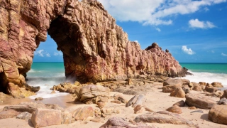 Formação rochosa na praia, com uma abertura no meio, onde turistas costumam tirar fotos