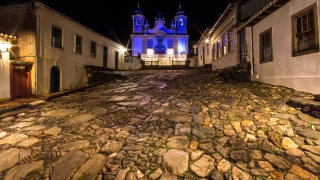 Vista noturna de igreja colonial em estilo barroco em cidade histórica de Minas Gerais. No plano frontal, destaque para a rua de pedra