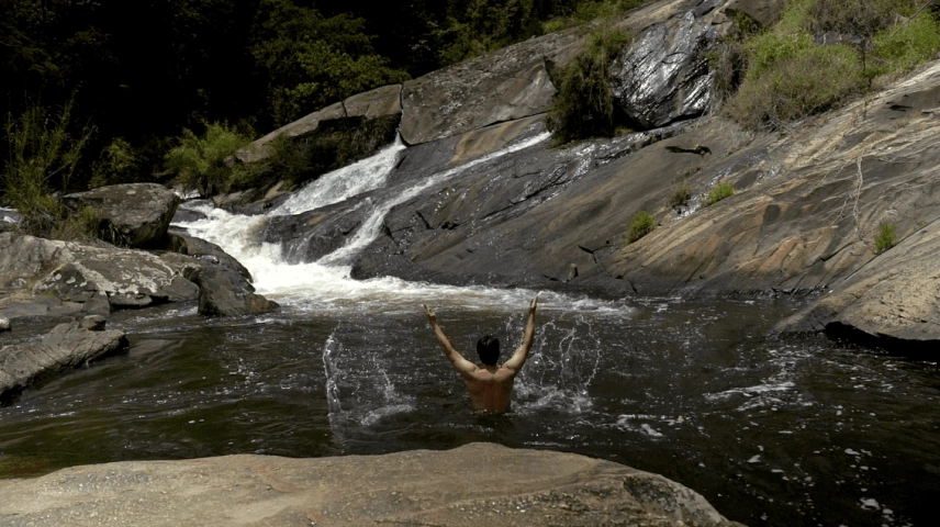 Homem de costas com os braços abertos em uma cachoeira, com rochas aos lados