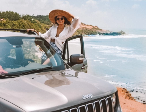 Mulher usando chapéu e roupas tropicais enquanto faz pose para foto na porta do carro que está estacionado em uma praia. Céu azul com poucas nuvens ao fundo. A foto serve de ilustração para um artigo sobre o tema “quanto custa alugar um carro”.