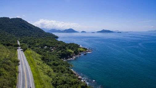 Vista aérea de estrada asfaltada ao lado esquerdo da imagem, vegetação ao centro e mar azulado à direita