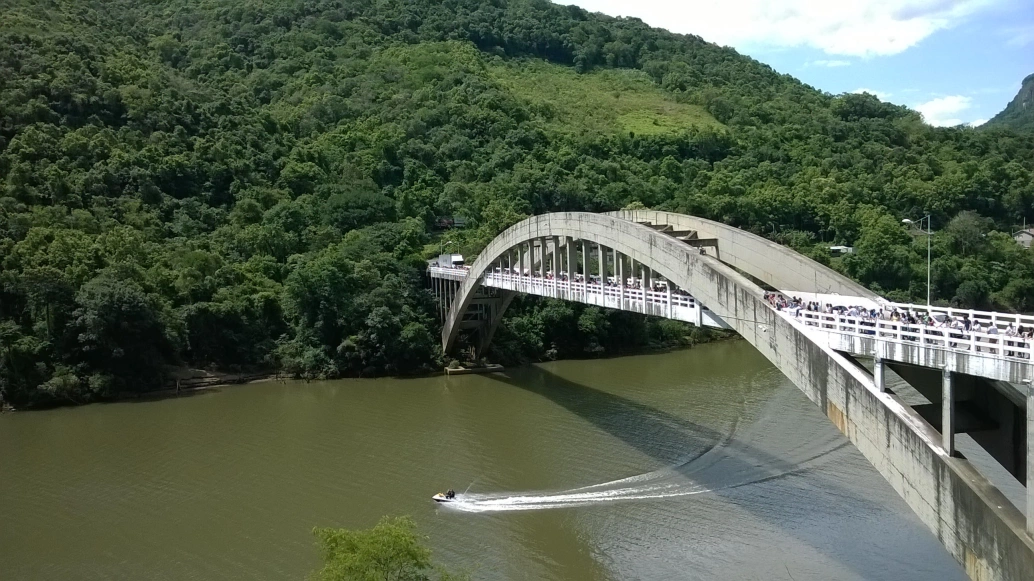Vista aérea de ponte com dois enormes arcos paralelos ao longo de sua extensão. A construção passa por cima de uma grande rio e, no final, há uma colina com cobertura vegetal