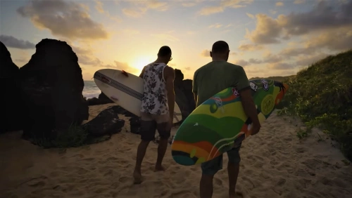 Dois homens caminham em praia segurando pranchas de surfe