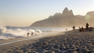 Pessoas na beira da praia de Ipanema aproveitando dia ensolarado e ao fundo a paisagem do Morro Dois Irmãos