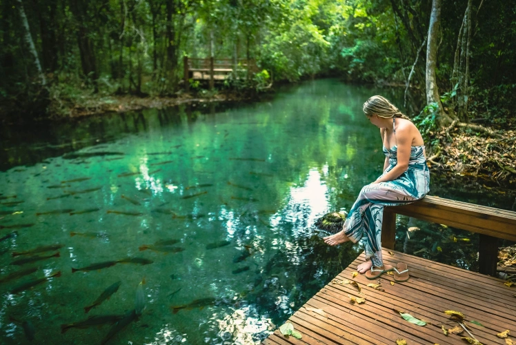 Mulher sentada em banco de madeira contempla peixes em lagoa de água azul cristalina em Nobres MT