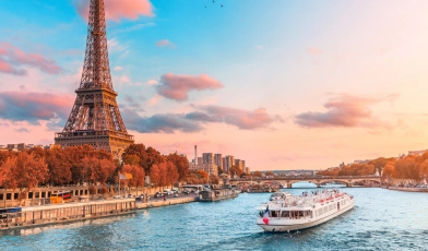 Fotografia da Torre Eiffel ao lado do Rio Sena, por onde passa um barco branco. Céu azul turquesa com um pequeno grupo de pássaros e algumas nuvens.