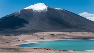 Laguna verde, com águas cor de esmeralda, envolta de terreno baixo e árido. Ao fundo, um imponente e gigante vulcão, Ojos del Salado