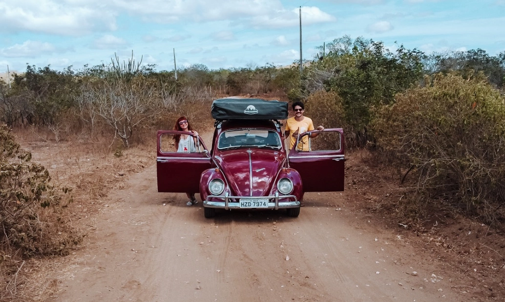 Um carro modelo Fusca de cor vermelha parado em estrada de terra em dia claro com um homem e uma mulher em pé apoiados em suas portas