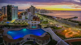 Vista aérea de cenário urbano em contraste com o pôr do sol refletindo na praia. No plano frontal, uma enorme piscina com as bordas curvadas e prédios ao fundo